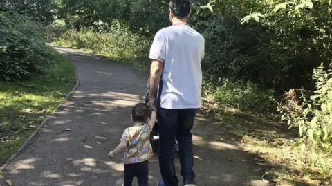 A Syrian asylum seeker, whose face we cannot see, walks down a path outside with his child.