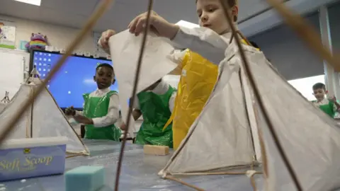 One child holds up paper for a lantern they are making in a classroom 