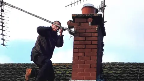 Dennis Iliffe A man in black clothing and brown boots kneels on a tile roof, working on an aerial which is attached to a chimney.