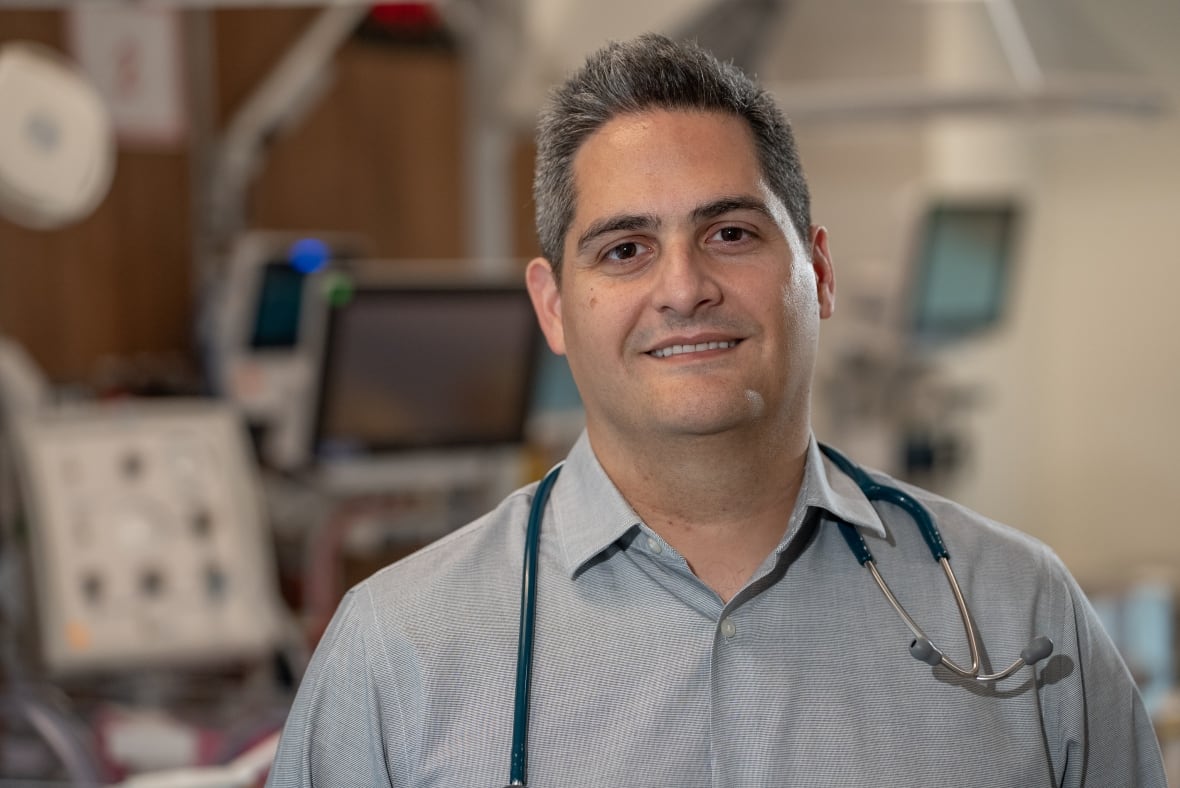 A man smiling and wearing a stethoscope with medical equipment behind him.