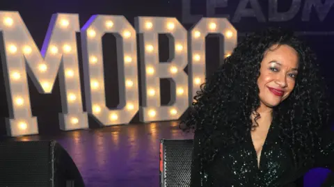 Getty Images Kanya King at the 2024 MOBO Awards. Kanya has her hand on her hip and smiles at the camera, wearing a sparkly black dress. Behind her is a stage with sound equipment and MOBO spelled out in big lights. 