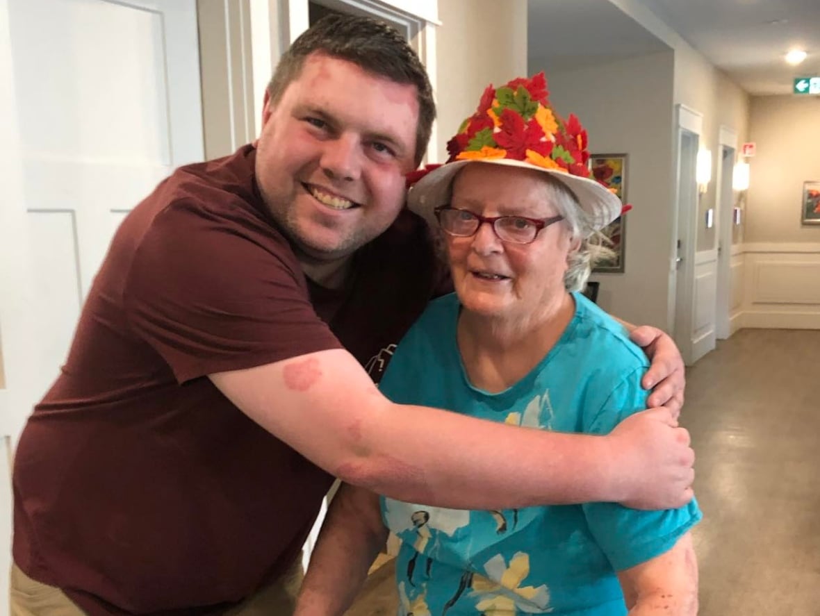 A white man wearing maroon t-shirt is hugging a shorter, older woman, wearing a turqoise shirt and a colourful hat. They are standing in a hallway. She is holding onto a walker.