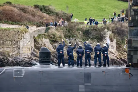 Royal Navy The submarine arriving at port, with five personell on deck in white hats seen from the back waving to people on a rocky and grassy shoreline.