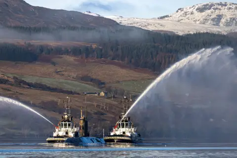 Royal Navy HMS Triumph submarine  at sea, with two tugboats sailing alongside it and firing their water cannons. Hills and trees are visible in the background. 