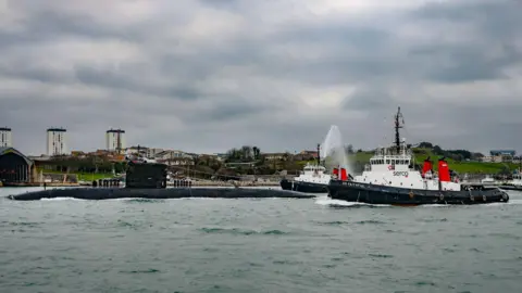 Royal Navy The submarine arriving at port, accompanied by two tugboats firing water cannons. The skies above are grey and overcast