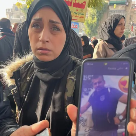 A woman in the crowds outside the hospital holds up her phone showing a photo of the relative she's looking for