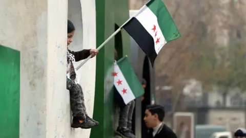 A child sits on a window sill waving a new Syria flag.