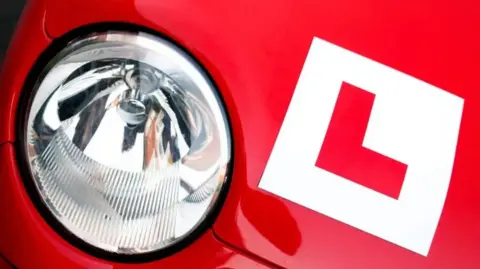 Getty Images Red Car and its headlight with a learner plate near it, which is a red L in a white background