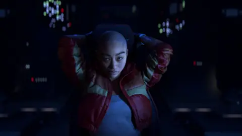 Naughty Dog A woman with a shaved head wearing a red leather jacket sits in a darkened room with both hands placed behind her head. She's illuminated by lights from nearby control panels and screens.