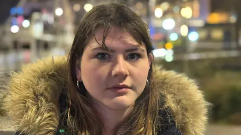 A woman with long brown hair, earrings and wearing a coat with a furry hood.