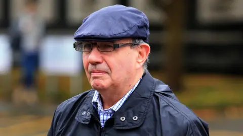 PA Media A man with black-rimmed glasses, wearing a blue flat cap, a blue jacket and a blue and white checked shirt underneath. He is walking into court, but only his shoulders and face can be seen in this image.