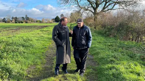 Peter Garsden, a man wearing a long wool coat and with glasses, talks with his client Keith Levell in a field. Keith has dark trousers, a dark coat and a baseball cap.