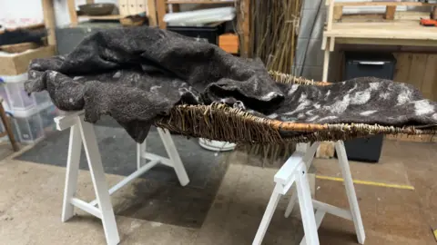 Lizzy Steel / BBC A woollen, black-and-white burial shroud sits in a wicker basket in an artist workshop. It is displayed on top of a white wooden stand. 