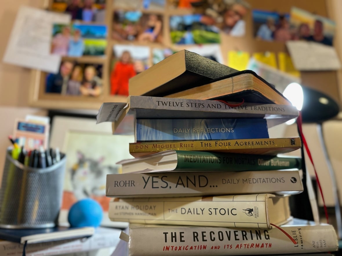 A stack of books on a table. In the background is a corkboard with several pinned photos.