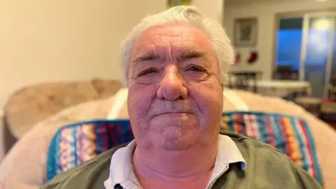 A man with white hair and dark bushy eyebrows sits in a living room and smiles. There is a window behind him and it is nearly dark outside.