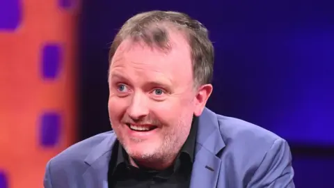 PA Media Chris McCausland pictured smiling and wearing a blue jacket and black shirt on a TV studio set.