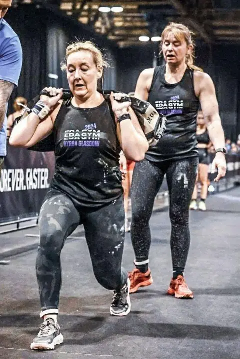 Hyrox and Sportograf Leigh completes a lunge with a weighted sand bag on her back. Her race partner Wiki follows behind her waiting for her turn