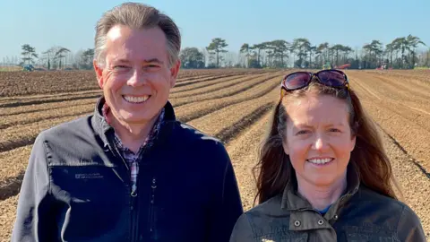 Tracy Bush A middle-aged man and woman are stood next to each other in the foreground of the picture. The man has short grey hair and is wearing a jacket over a check shirt. To his left is the woman, with sunglasses on her head and long brown hair, also wearing a jacket. Both are smiling and looking at the camera. In the background is a ploughed field, with agricultural machinery and a line of trees visible in the distance. The sky is clear and blue.