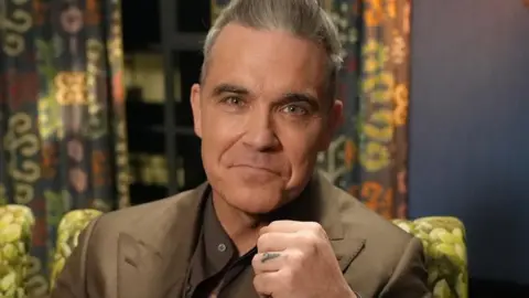 Robbie Williams pictured smiling whilst raising a fist to the camera