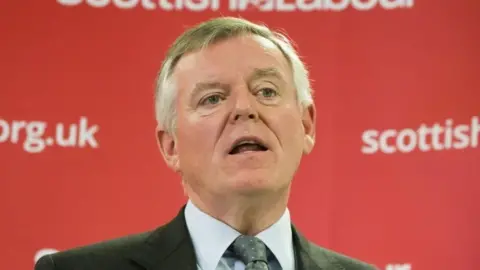 PA Media Graeme Pearson photographed against a red backdrop with the Scottish Labour website details in white writing. His mouth is open as if he is speaking. Pearson, who has grey hair combed in a side shed, is wearing a grey suit, light grey shirt and dark grey tie.