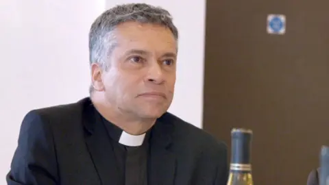 Picture of David Tudor who has short greying hair and is wearing a black clerical shirt with white clerical collar. He is pictured sitting down with a wall and a brown door behind him.