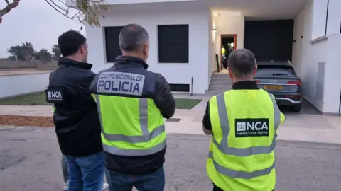 NCA One Spanish Policeman and two NCA officers wearing hi vis at Colin Wright's home in Spain. a white building with one car outside.