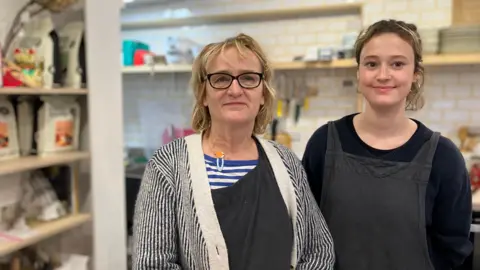 Justine Hyde with a member of staff at Hyde and Seek in Exeter, she wears a blue and white striped top with a hair clip on the collar, a black apron and a navy and white striped cardigan, her colleague is dressed in black with a black apron. 