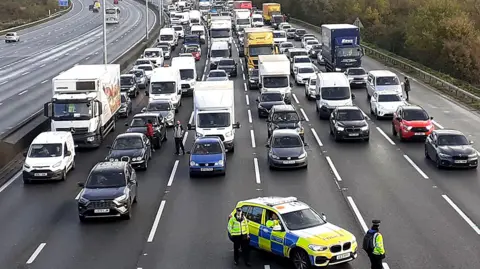 Just Stop Oil / PA Traffic stopped on M25, lined up like in a car park, with some people standing outside their cars, and a police car with three officers