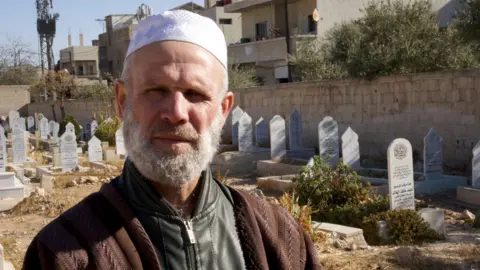 BBC/Thanyarat Doksone Abdul Kader al-Sheikha, a Muslim cleric, stands in Qutayfah cemetery