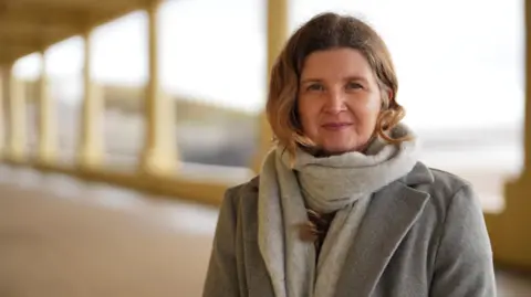 Johanna Robinson faces the camera with a neutral expression. She has dark blonde hair, and is outside on a costal path, wearing a grey coat and grey scarf. 