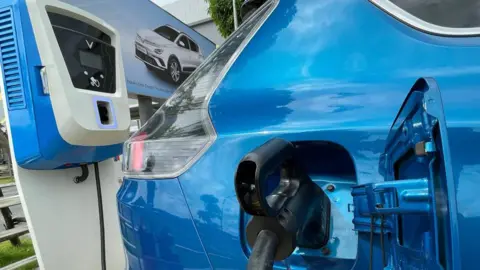 Getty Images A bright blue VinFast electric car plugged in at an outdoor charging station. Only the rear of the vehicle can be seen, with the charging hatch open. A picture of a grey version of the vehicle can be seen next to the car.