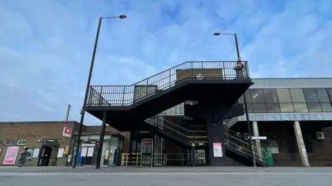 Ben Schofield/BBC The exterior of Luton Station, as seen from the town centre side of the tracks. Two lampposts stand either side of a set of stairs that dog-leg from the street level to an un-seen ticket office. A red and white railway sign can be seen on the left of the image. There is  tarmac in the foreground and a blue sky with thin clouds hangs above the station.