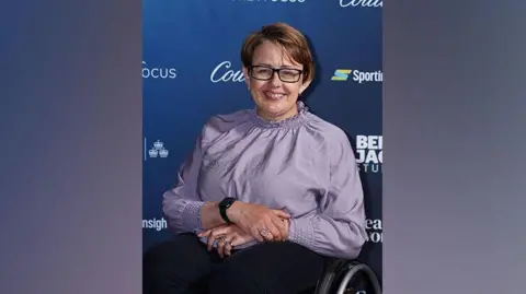 PA Media Baroness Tanni Grey-Thompson looking direct to camera. She is wearing a lilac top, with frilled collar and cuffs, black trousers, and dark framed, rectangular glasses. She is seated in a wheelchair. The top of one wheel is visible on the right of the image. Her hands are folded on her lap. She is posing in front of an advertising hoarding, which has the names of several brands printed on it.