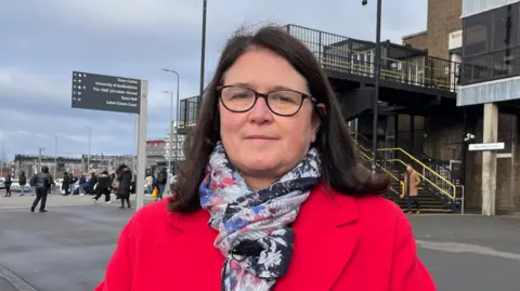 Ben Schofield/BBC Rachel Hopkins, wearing a bright red overcoat and patterned silk scarf, is standing outside Luton Station looking direct to camera. She is wearing dark rimmed glasses and has brown, shoulder-length hair. Behind her, the station entrance is seen, with pedestrians milling about and some signs.