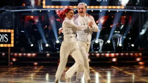 Chris McCausland and Dianne Buswell in matching white suits, in the middle of a dance in the Strictly final