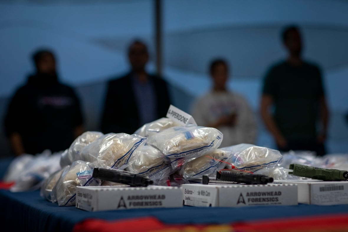 four figures, nearly silhouettes, stand in the background behind a pile of bagged drugs on a table. There are three handguns on white cardboard boxes that say "arrowhead forensics" on them in the foreground.