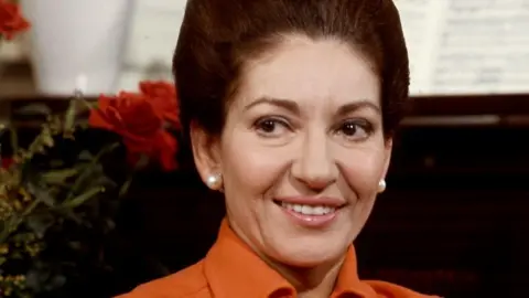 A picture of Maria Callas with her brown hair up, wearing pearl stud earrings and an orange shirt