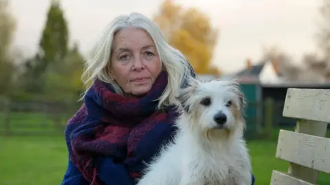 Lisa Willis, a woman with pale blond hair, wrapped in a large blue and purple scarf and a deep blue coat, sitting outside on a bench with grass, trees and a handful of rooftops visible behind her. Duke, her terrier, who has white fur with grey and brown patches around his ears, eyes and muzzle, is sitting beside her.