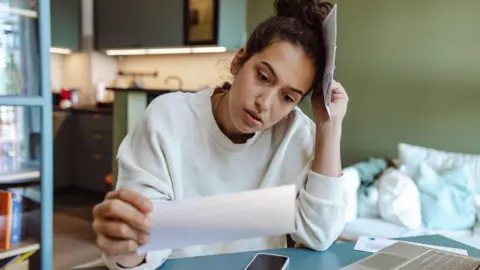 Getty Images A woman wearing a white jumper sits at a dining table while looking at her water bill.