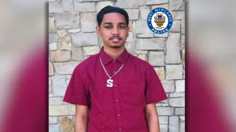 Family Handout A young man with dark hair and eyebrows, a sparse moustache and goatee beard. He is wearing a maroon short-sleeved shirt and standing in front of a stone wall.