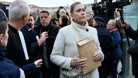 Getty Images Caroline Darian, wearing a cream coat, walks through a crowd, holding a brown envelope. She is followed by her brothers and there are cameras to her left.