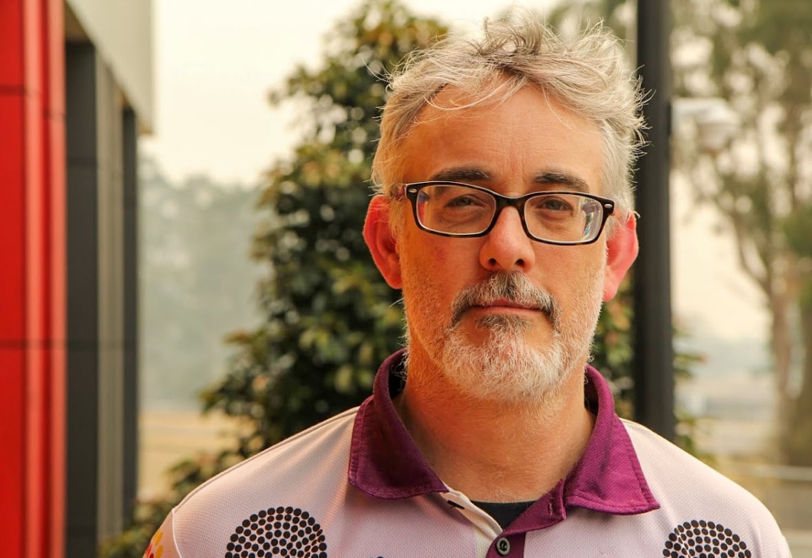 A man with grey hair, grey beard and black rectangular glasses.