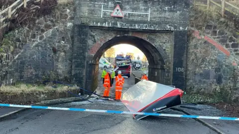 a bus roof sits on the ground under a bridge. A police cordon is visible in the foreground and emergency service workers wearing orange can be seen.