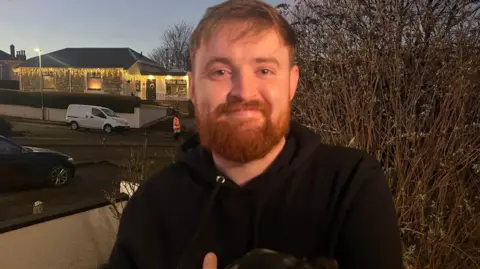 A man with orange hair and a beard wearing a black hoodie. He is standing in front of a hedge.