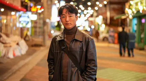 BBC/Maxine Collins Ryu Seonghyun standing on a street in a brown leather jacket, brown shirt and pants.