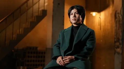 BBC/Hosu Lee Haneul sitting on a chair in a dimly-lit room wearing a dark green coat while he speaks to the BBC.