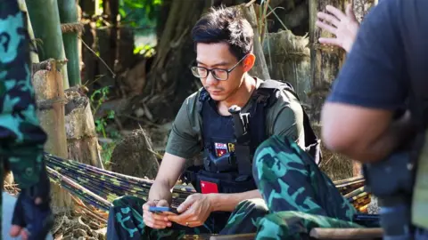 Ko BC / BBC Daeva, wearing a khaki t-shirt and military-style vest, is sitting on a camo-patterned hammock in the forest looking at his phone