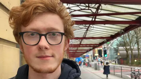 A close-up of Daniel, a young ginger-haired man with facial hair, looking at the camera with a neutral expression. He wears dark-framed glasses and a dark hoodie. Stretching into the distance above him is the metal and glass canopy sticking out from a train station wall. Blurred to the right of him is a section of pavement, railings and road, with a number of pedestrians walking along.