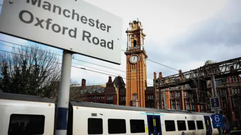 EPA-EFE/REX/Shutterstock Sign saying 'Manchester Oxford Road' in front of train and tall, narrow clock tower at Manchester Oxford Road station.