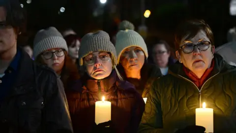 Pacemaker A close-up of four women and one man, who is partially obscured at the vigil. Three of the women are wearing woolen hats and three are also wearing glasses. Two of them are holding large white candles